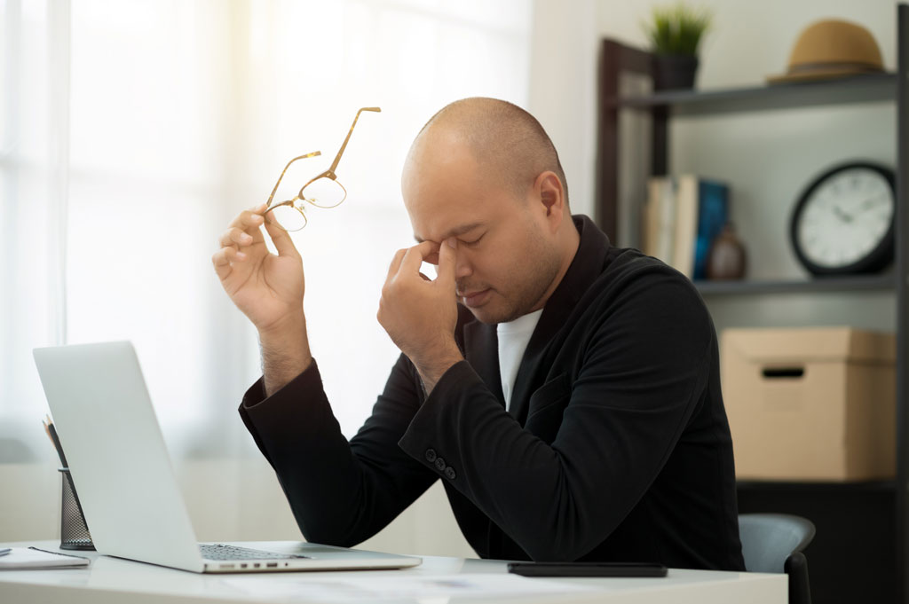 A man struggles with digital eye strain, sitting at a computer with his glasses off.